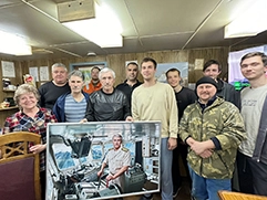 . На фото изображена группа людей, стоящих в помещении, которые улыбаются и позируют для снимка; в центре седой мужчина в черной одежде, он держит большую картину на холсте в раме, на которой он изображен в морской форме гражданского флота, стоящим за пультом управления судном; портрет выполнен в стиле под масло.