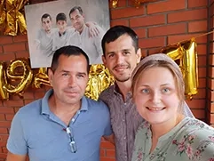 На фото изображена женщина и двое мужчин, они стоят рядом друг с другом и улыбаются; на заднем плане видна кирпичная стена, украшенная картиной на холсте, на которой изображены трое мужчин в белых рубашках; портрет выполнен в стиле под масло