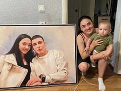 На фото женщина, держащая на руках ребенка, рядом с портретом на холсте в раме; на портрете изображены двое — она и её муж, они выглядят счастливыми и улыбаются, у женщины длинные темные волосы, а мужчина в светлой одежде; портрет выполнен в реалистичном стиле