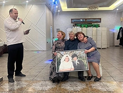 Фото с изображением пожилой пары и их дочери; они сидят и держат в руках картину на холсте в раме в стиле под масло; они находятся в банкетном зале или кафе; сбоку мужчина с микрофоном; атмосфера выглядит праздничной, с декоративным освещением и зеркалами на стенах; на картине пожилая пара нарисована по старому фото; на ней они молоды и только поженились.