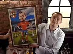 На фото изображён улыбающийся человек в белой рубашке, который держит шарж; на шарже — футболист с его лицом в красно-синей форме с надписью московского «ЦСКА» на груди; футболист стоит на футбольном поле рядом с воротами, а на заднем плане виден стадион