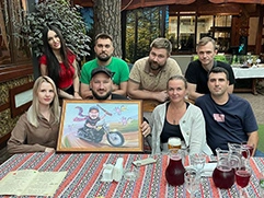 На фото группа людей, сидящих за столом на уличной веранде ресторана; на столе стоят кувшины с напитком, стаканы, меню и другие предметы; в центре стола сидит именинник с подарком — шаржем на холсте, он изображён с ружьём на мотоцикле, окружённый яркими элементами, такими как птицы и цветы