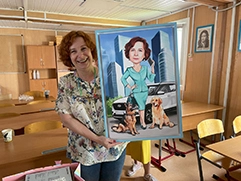 На фото женщина, которая стоит в классе, улыбается и держит в руках подарок — шарж на холсте в раме; на шарже нарисована она сама в деловом костюме, стоящая рядом с двумя собаками — немецкой овчаркой и золотистым ретривером; на заднем плане шаржа виден городской пейзаж с небоскребами и автомобилем